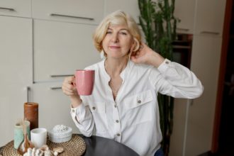 50-year-old blonde woman holding cup of tea and sitting at modern kitchen.