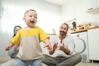 Caucasian baby boy child learn to walk with parents support to develop skill in living room at home.